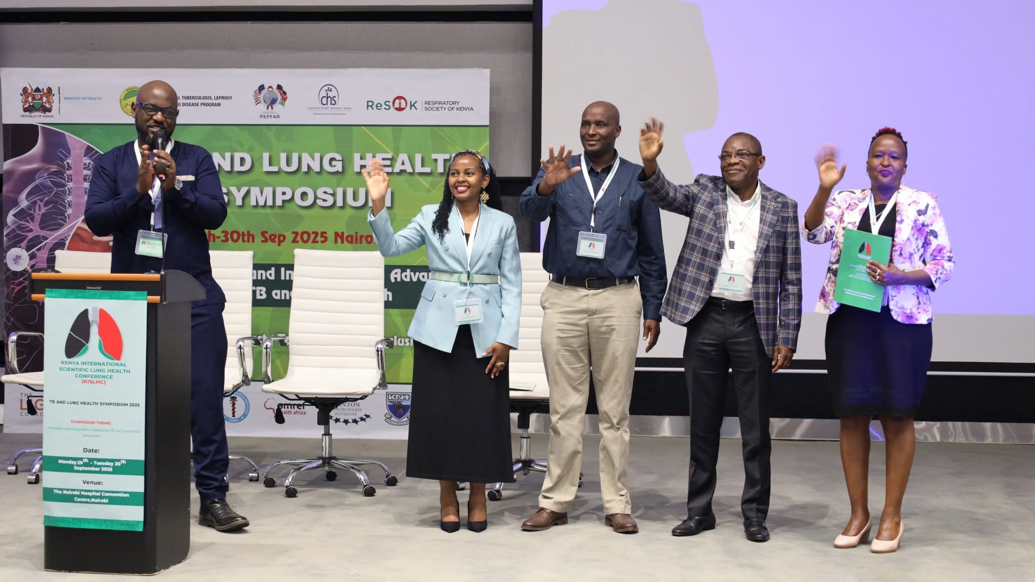 Speakers and guests waving on stage beneath the symposium banner